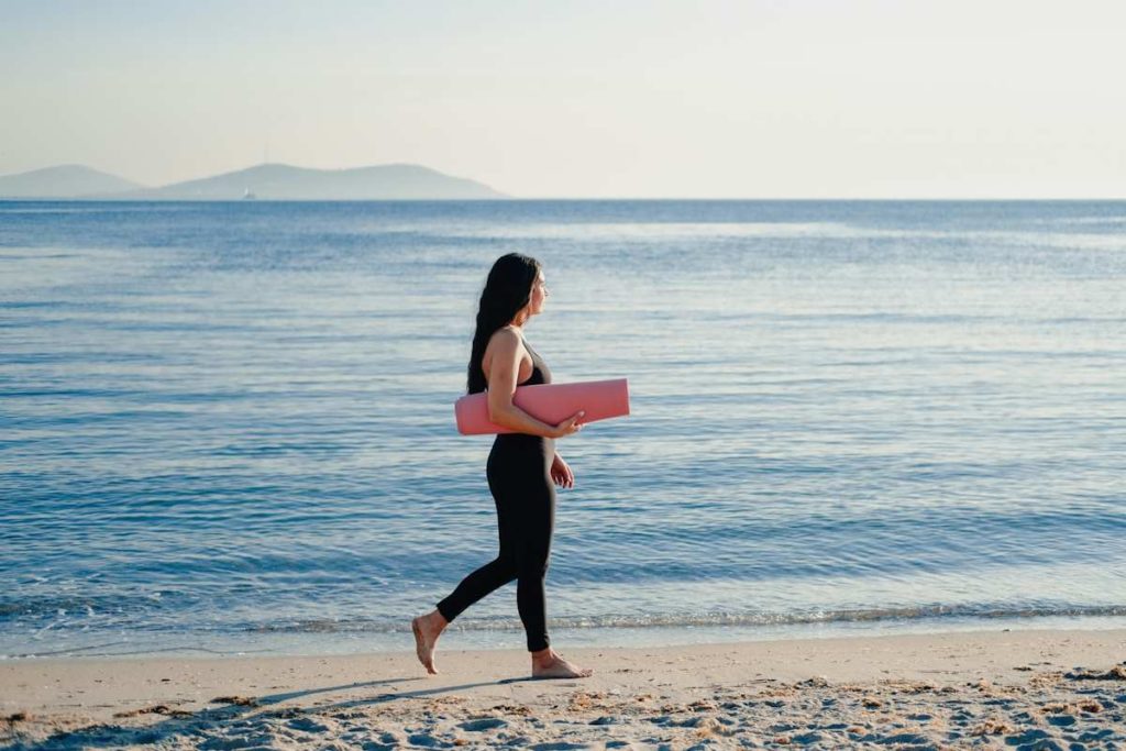 Une dame marche sur le sable avec son tapis de yoga.