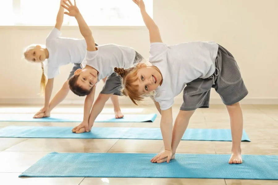 Trois enfants suivent une leçon de yoga pour enfant sur un tapis bleu.
